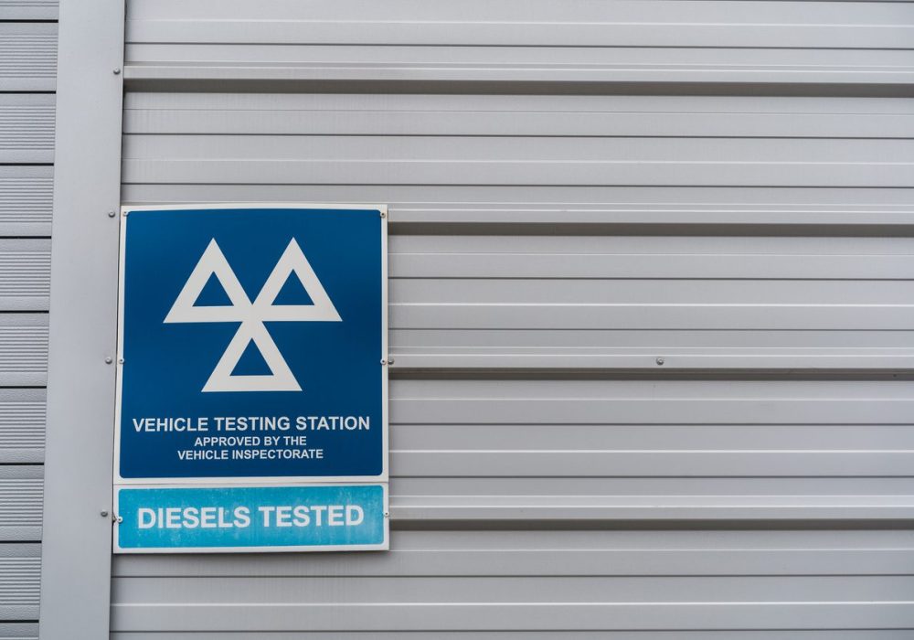 A blue vehicle testing station MOT sign, against a gray shuttered wall.